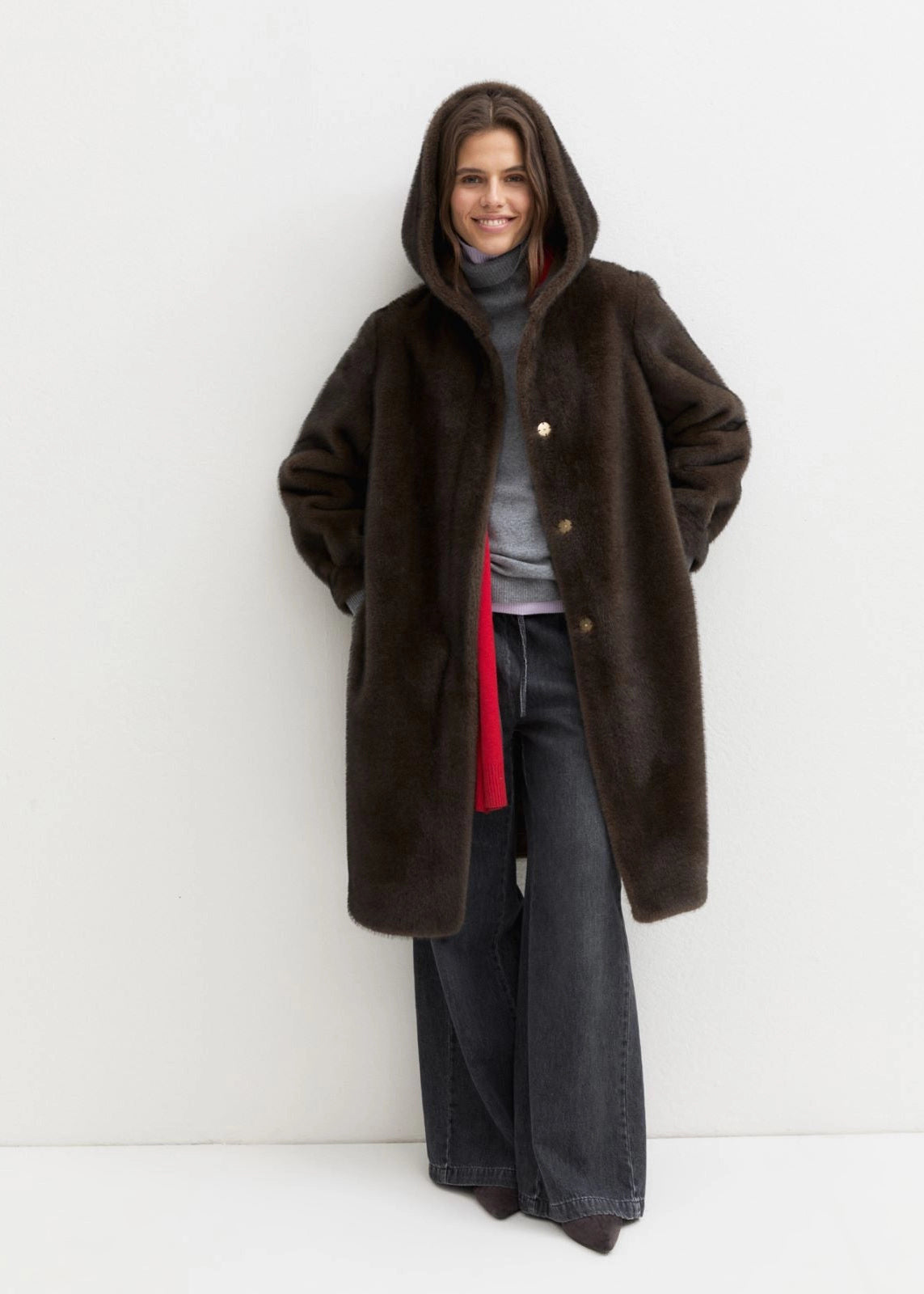 A woman wearing a dark brown hooded faux fur coat over a gray turtleneck, a red shirt, and wide-leg dark jeans, standing against a plain white studio background.