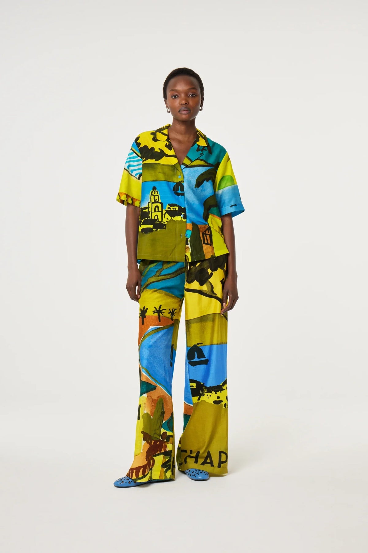 A model stands against a light gray studio background wearing a matching short-sleeve button-up shirt and wide-leg trousers set in a bold multicolor landscape print featuring blue, yellow, green, and orange.