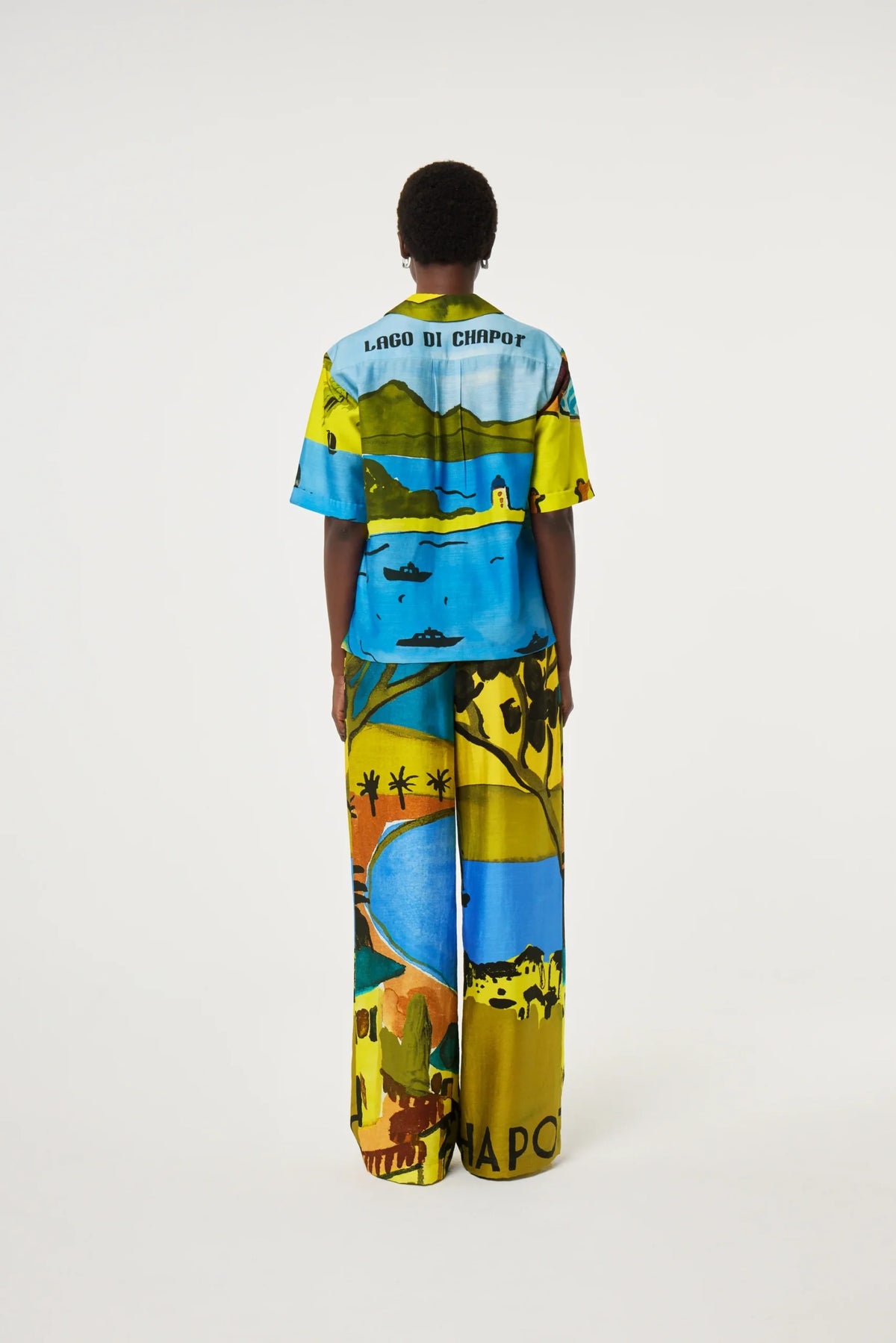 A model viewed from the back wearing a matching short-sleeve t-shirt and wide-leg trousers set with a vibrant, multicolor landscape print featuring blue water, green hills, and yellow fields. The shirt has 'LAGO DI CAMPO' printed on the upper back.