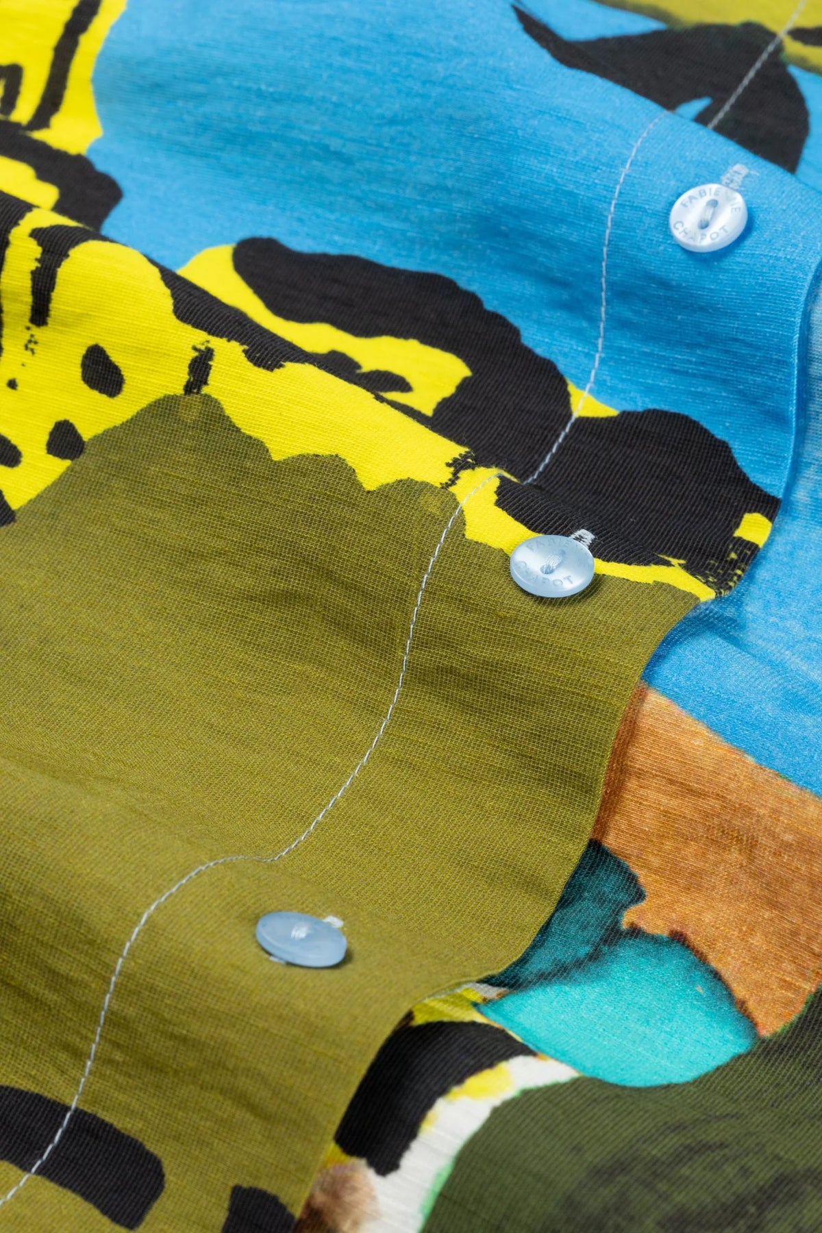 Close-up of a button-front garment with an abstract patchwork print in blue, olive green, yellow, black, and turquoise, shown against no visible background.