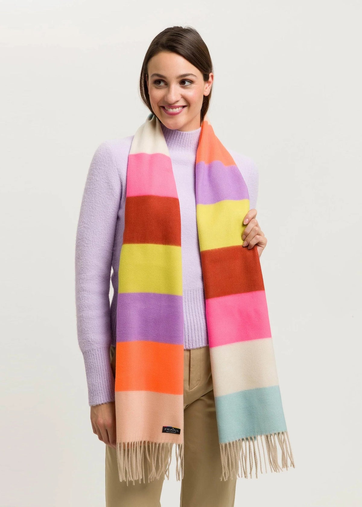 A woman wearing a pastel lavender sweater and beige pants models a thick striped scarf with fringe. The scarf features wide blocks of pink, orange, yellow, purple, red, and light blue, with a cream section at the top and bottom.