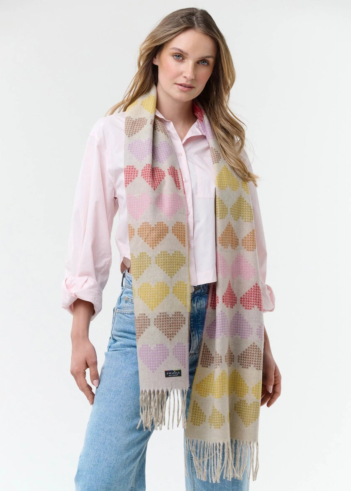 A woman modeling a cream-colored knit scarf with a repeating pattern of pastel pink, yellow, and purple hearts, styled over a light pink shirt and blue jeans against a white studio background.