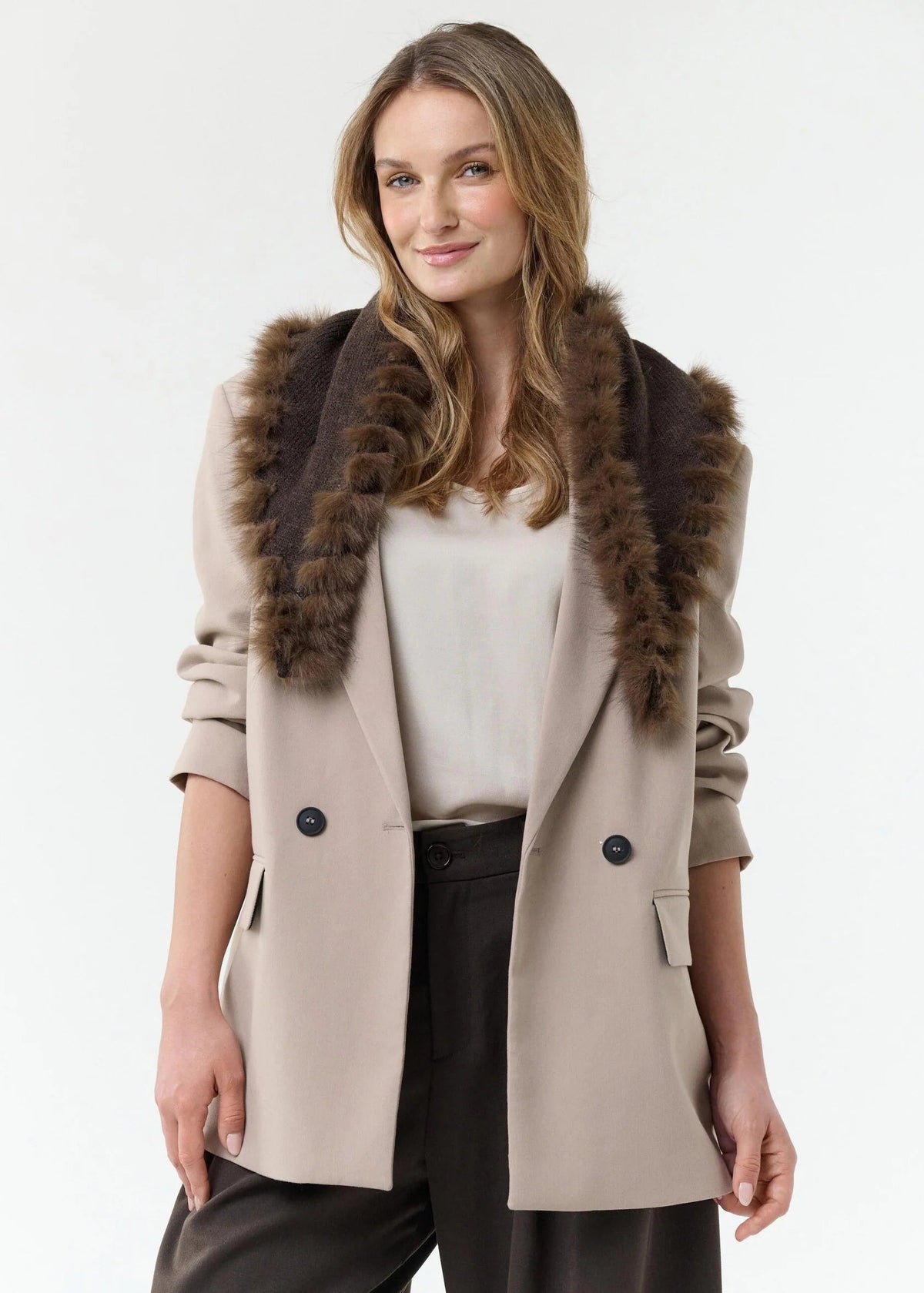 A woman wearing a beige double-breasted coat with a dark brown fur-trimmed collar, styled with a cream top and dark trousers, against a plain white studio background.