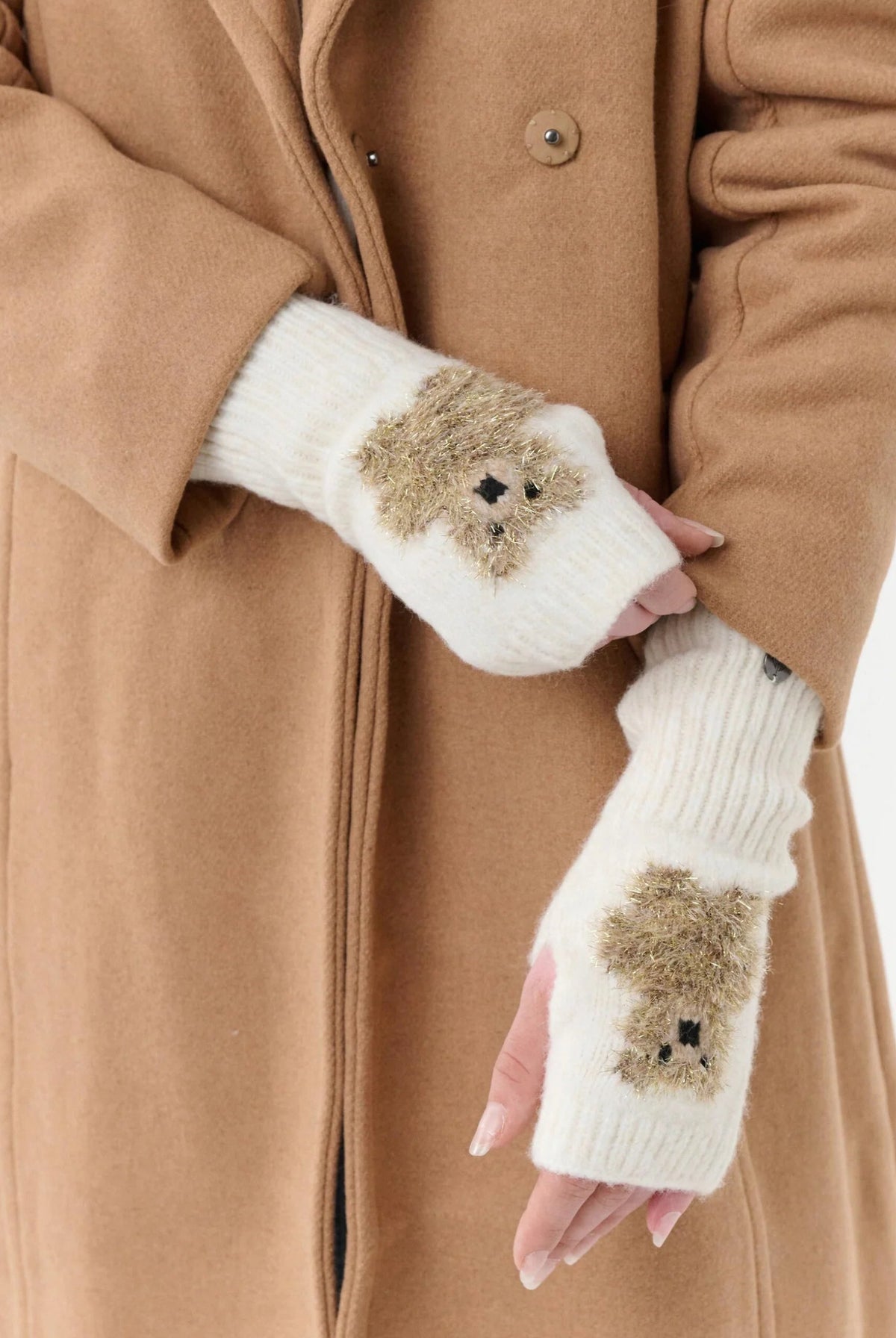 A person wearing a camel-colored coat and white fingerless knit gloves with small brown teddy bear faces on the palms, shown against a plain light background.