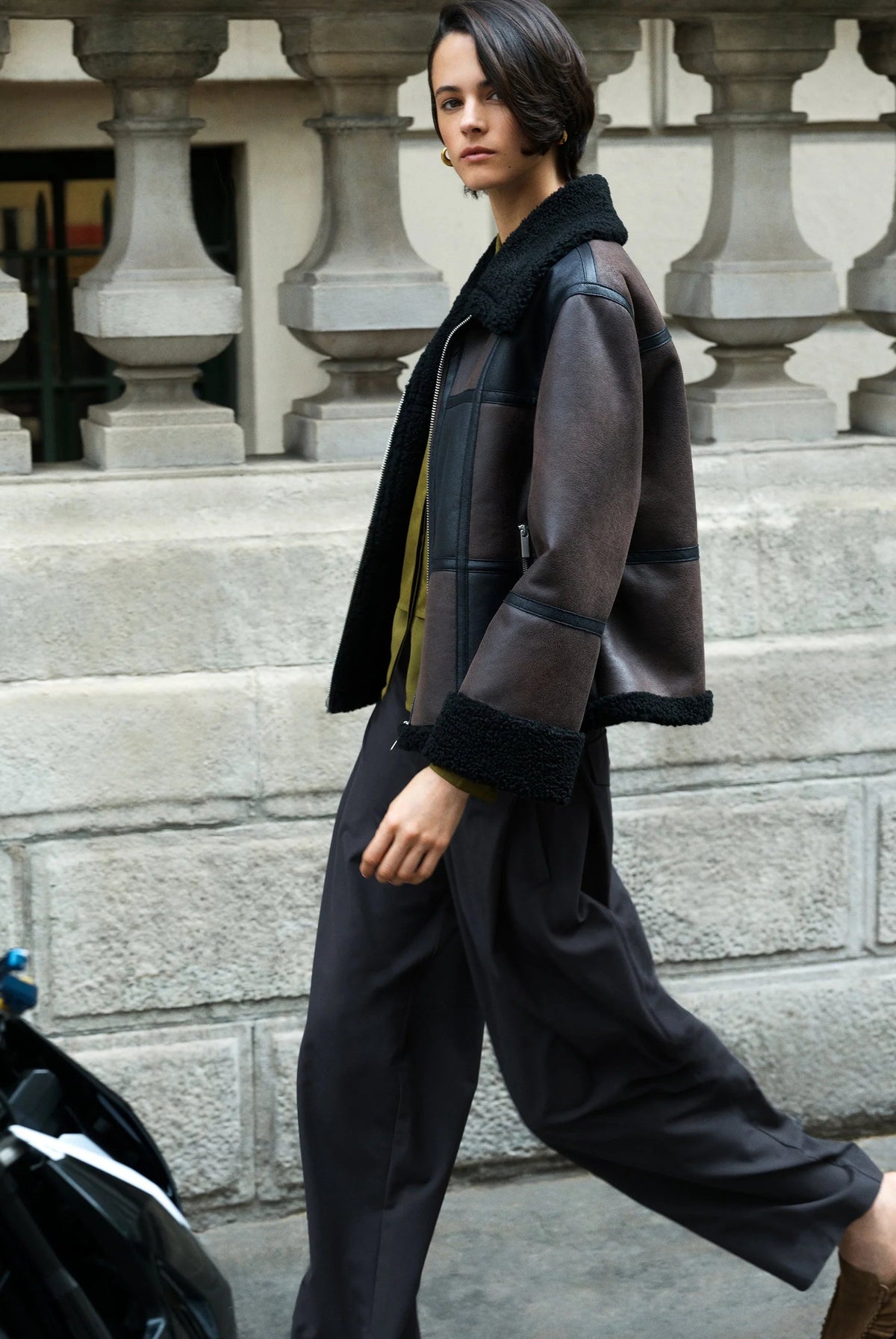 A woman walking outdoors in front of a stone balustrade, wearing a dark brown patchwork shearling jacket with black trim, black wide-leg trousers, and brown loafers.