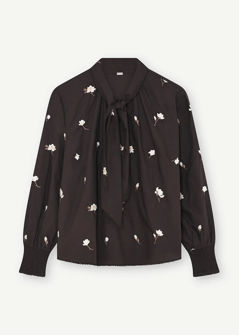 A black long-sleeve blouse with a small white floral print, featuring a tie-neck detail and a ruffled hem, shown against a light off-white background.