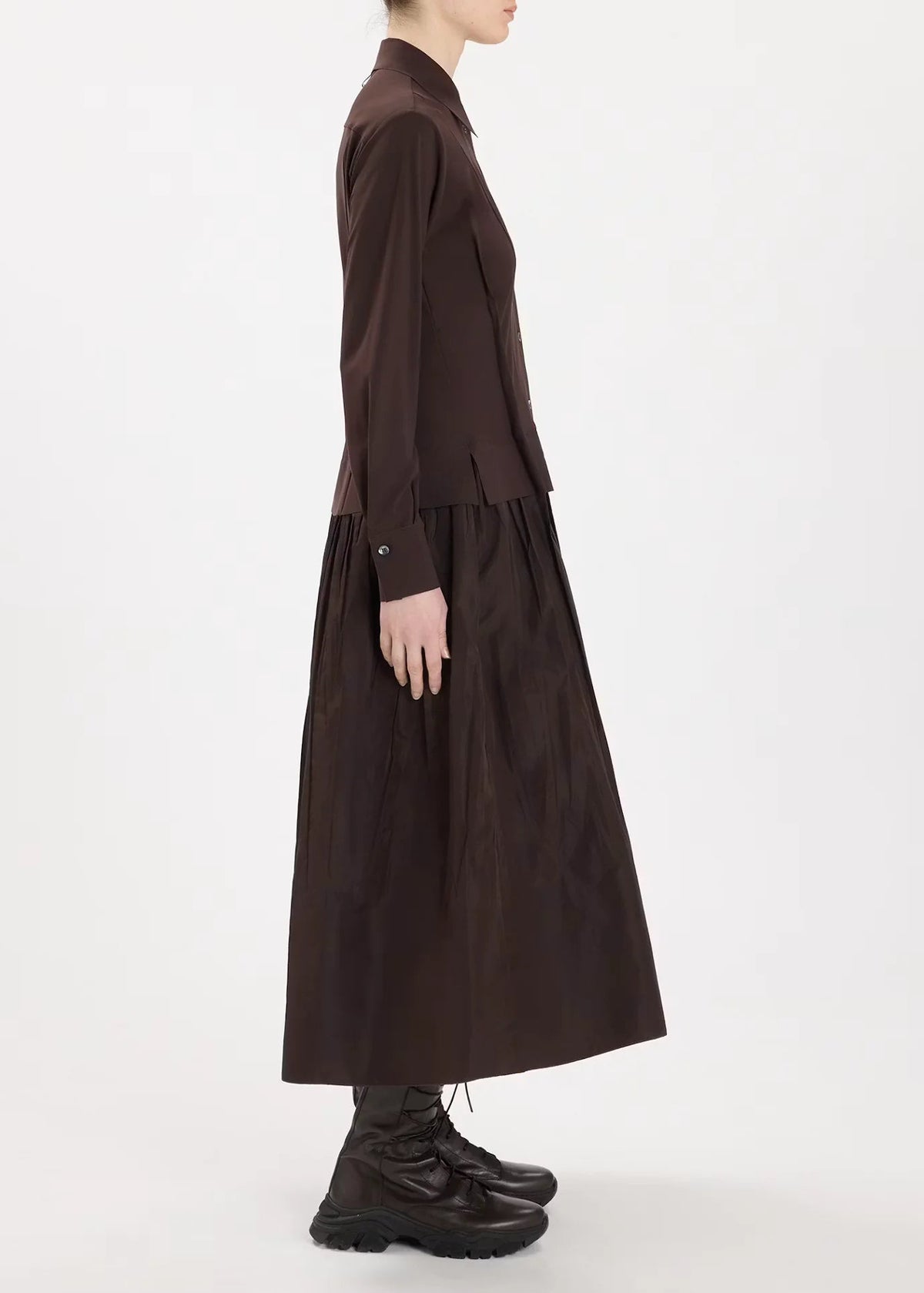 A model shown in profile wearing a dark brown long-sleeve collared top and matching pleated midi skirt, paired with dark brown lace-up boots, against a plain white studio background.