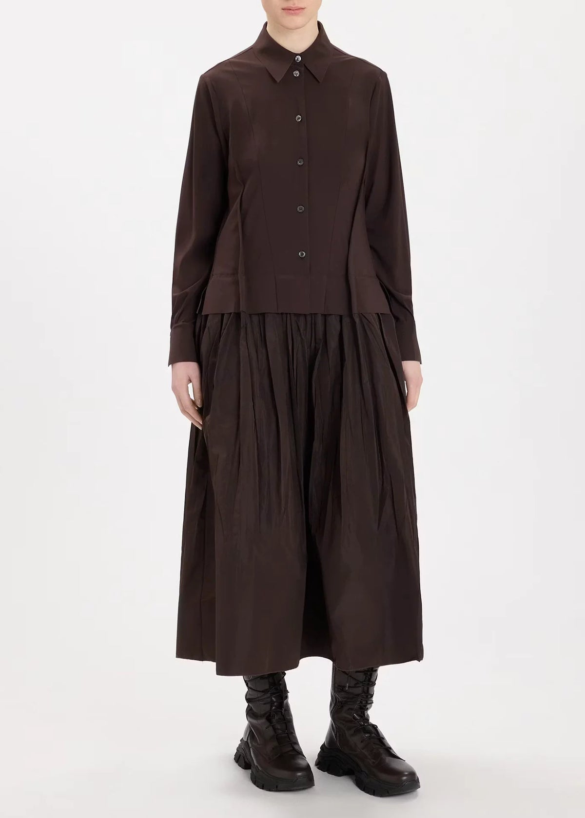 A model wearing a dark brown long-sleeve button-up shirt and matching pleated midi skirt, paired with black lace-up boots, against a plain white studio background.