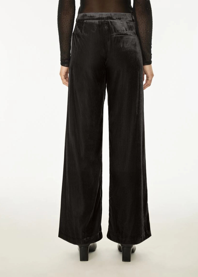 Black wide-leg velvet trousers shown from the back on a model, featuring a textured waistband and back pockets, paired with black heels.
