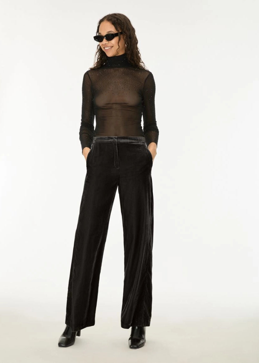 A model wearing a sheer black long-sleeve mock neck top and black wide-leg velvet trousers, styled with black sunglasses and black boots against a plain white studio background.
