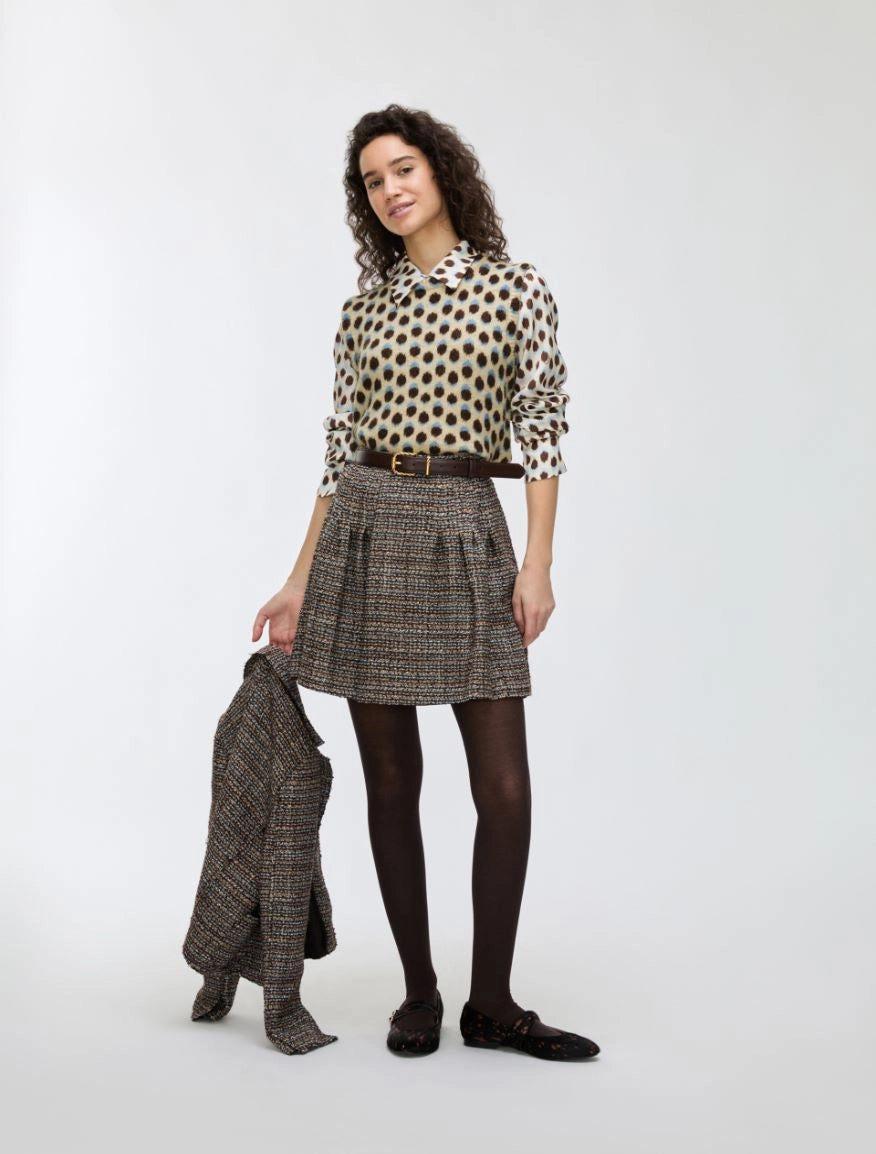 Woman wearing a polka dot blouse and patterned skirt holding a coat on a white background