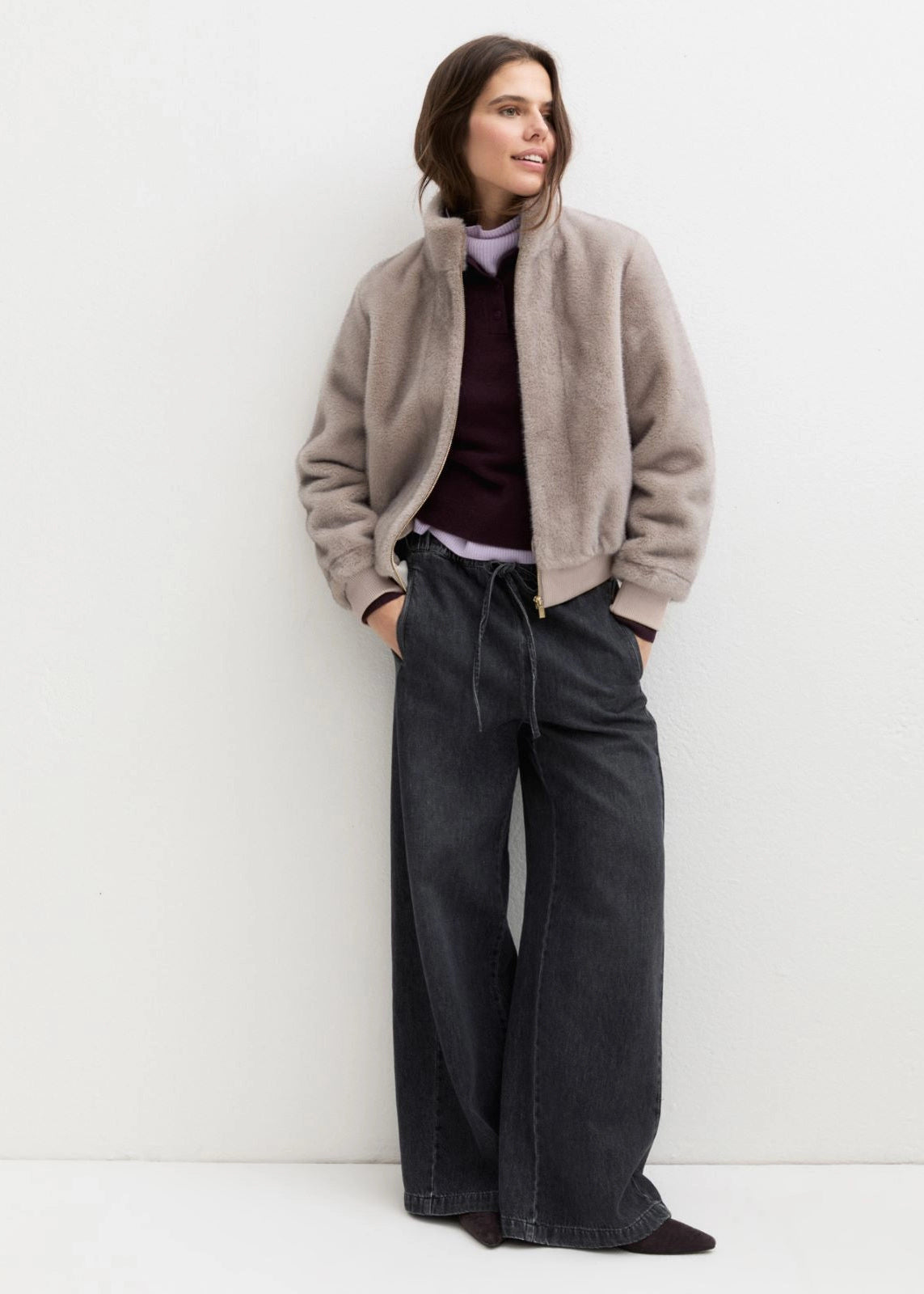 A woman stands against a plain light gray wall wearing a taupe fuzzy cropped jacket over a dark burgundy top and dark charcoal wide-leg jeans.