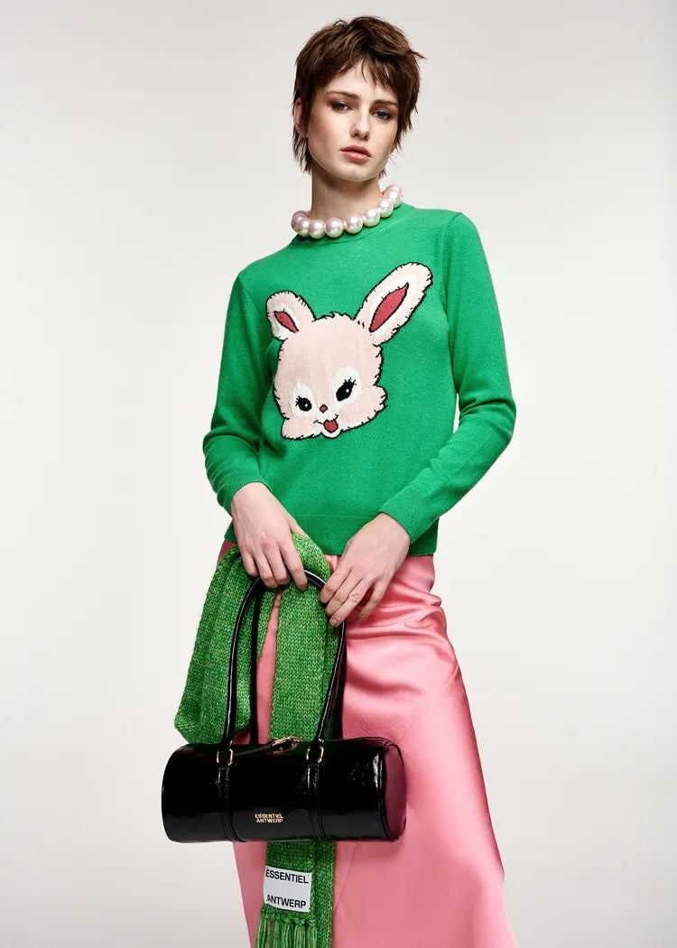 A model wearing a bright green knit sweater with a large white and pink bunny face graphic on the front, styled with a pink satin skirt, a green knit scarf, and a black cylindrical handbag against a light gray studio background.