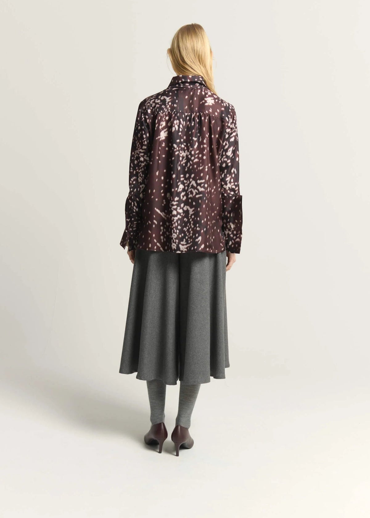 A woman stands with her back to the camera wearing a dark brown and black patterned long-sleeve shirt, a gray midi skirt, gray tights, and dark brown heels, against a plain light gray studio background.