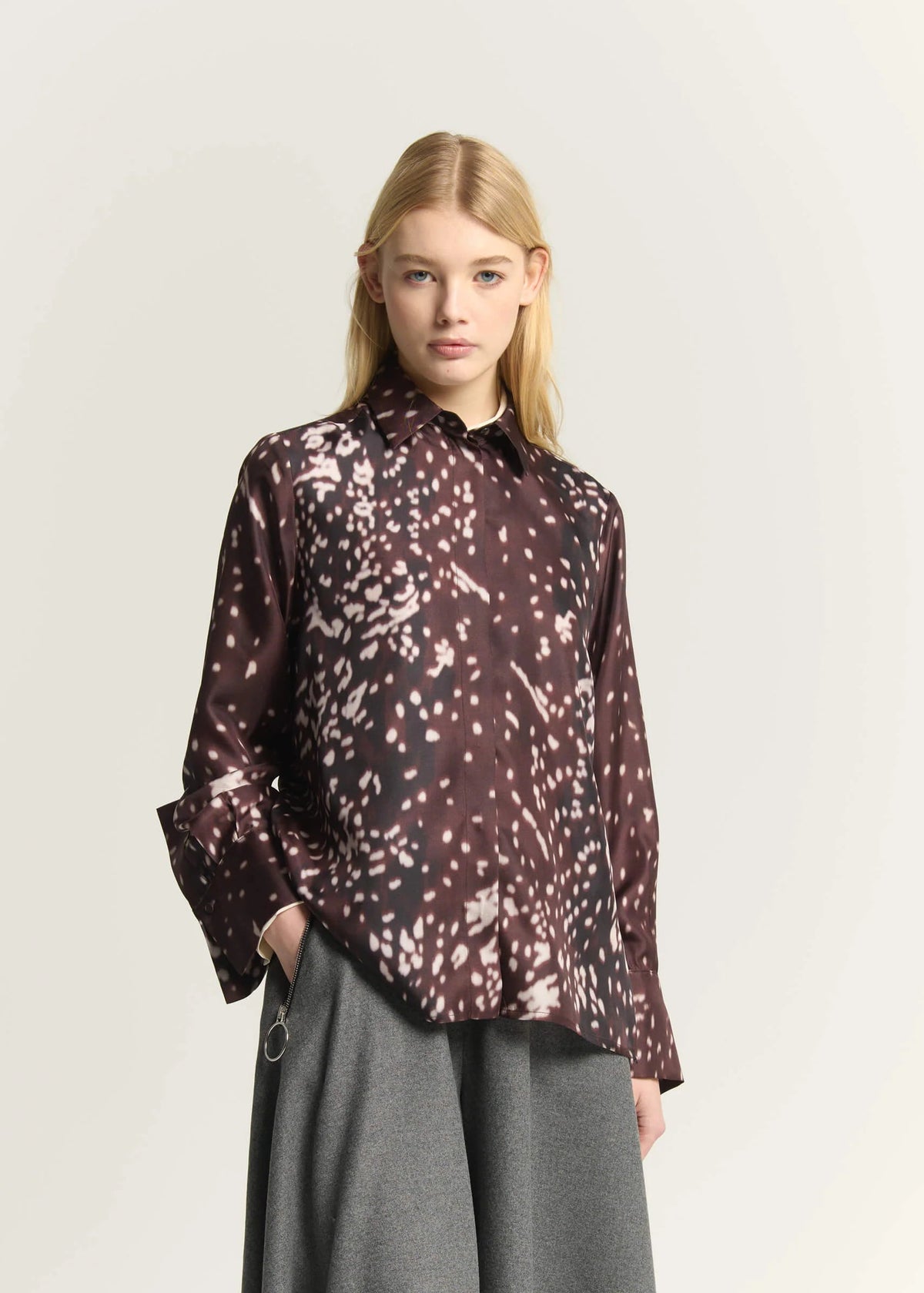 A model wearing a long-sleeve button-up shirt with a dark brown and black abstract spotted pattern, paired with gray wide-leg trousers, against a plain light gray background.