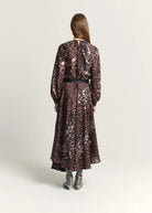 A woman viewed from the back wearing a long-sleeve midi dress with a dark brown and black abstract floral pattern, a black waist belt, and gray pointed-toe heels, set against a plain light gray studio background.