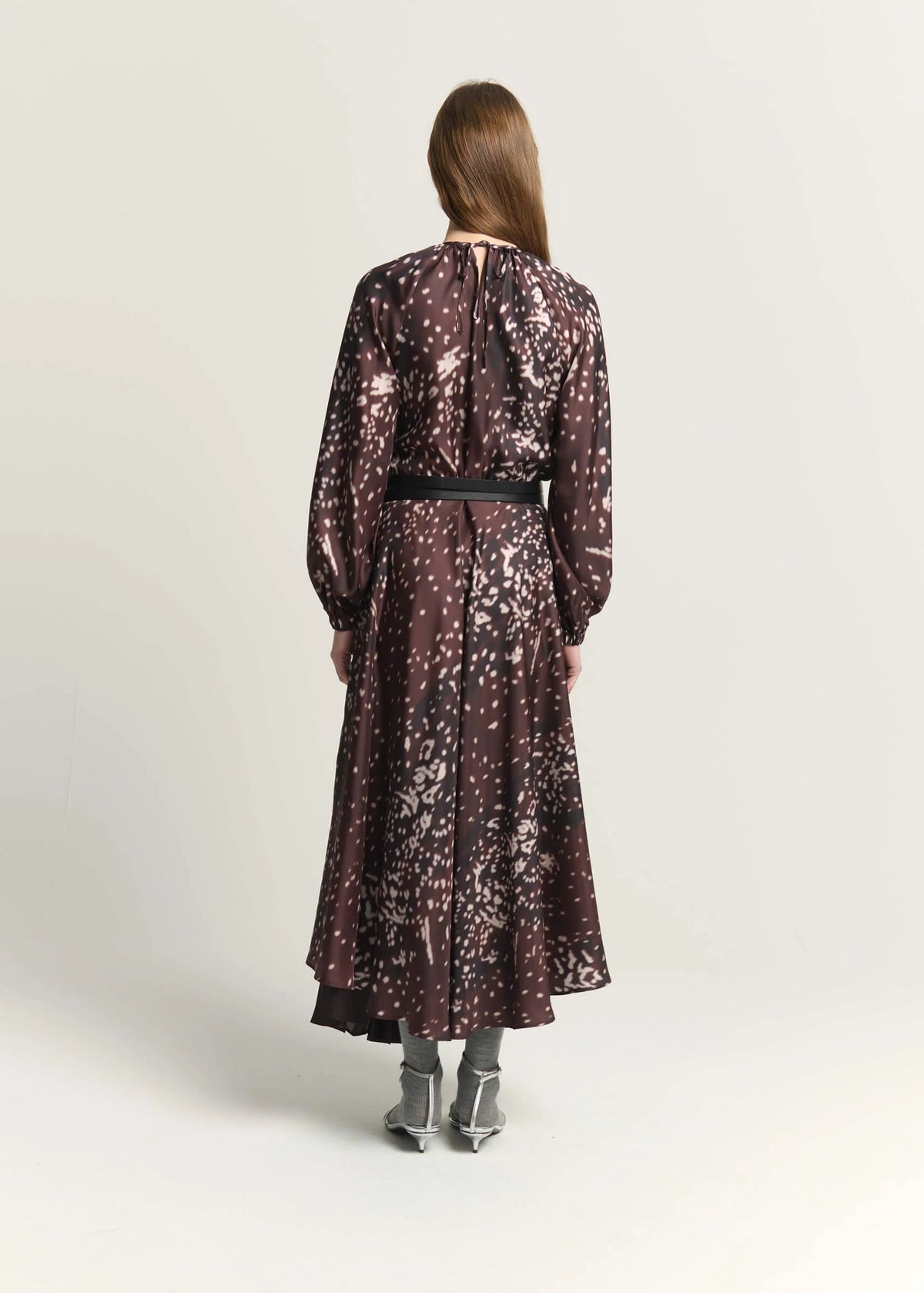 A woman viewed from the back wearing a long-sleeve midi dress with a dark brown and black abstract floral pattern, a black waist belt, and gray pointed-toe heels, set against a plain light gray studio background.