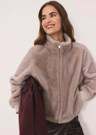 A woman wearing a soft, fuzzy high-neck zip-up jacket in a dusty taupe color, holding a dark burgundy coat under her arm against a plain white background.