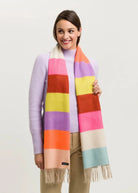 A woman wearing a pastel lavender sweater and beige pants models a thick striped scarf with fringe. The scarf features wide blocks of pink, orange, yellow, purple, red, and light blue, with a cream section at the top and bottom.