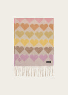 A rectangular woven scarf with fringe, featuring a repeating heart pattern in pink, orange, yellow, brown, and purple on a light beige background. A small black label with white text is visible near the bottom right corner.