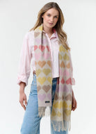 A woman modeling a cream-colored knit scarf with a repeating pattern of pastel pink, yellow, and purple hearts, styled over a light pink shirt and blue jeans against a white studio background.