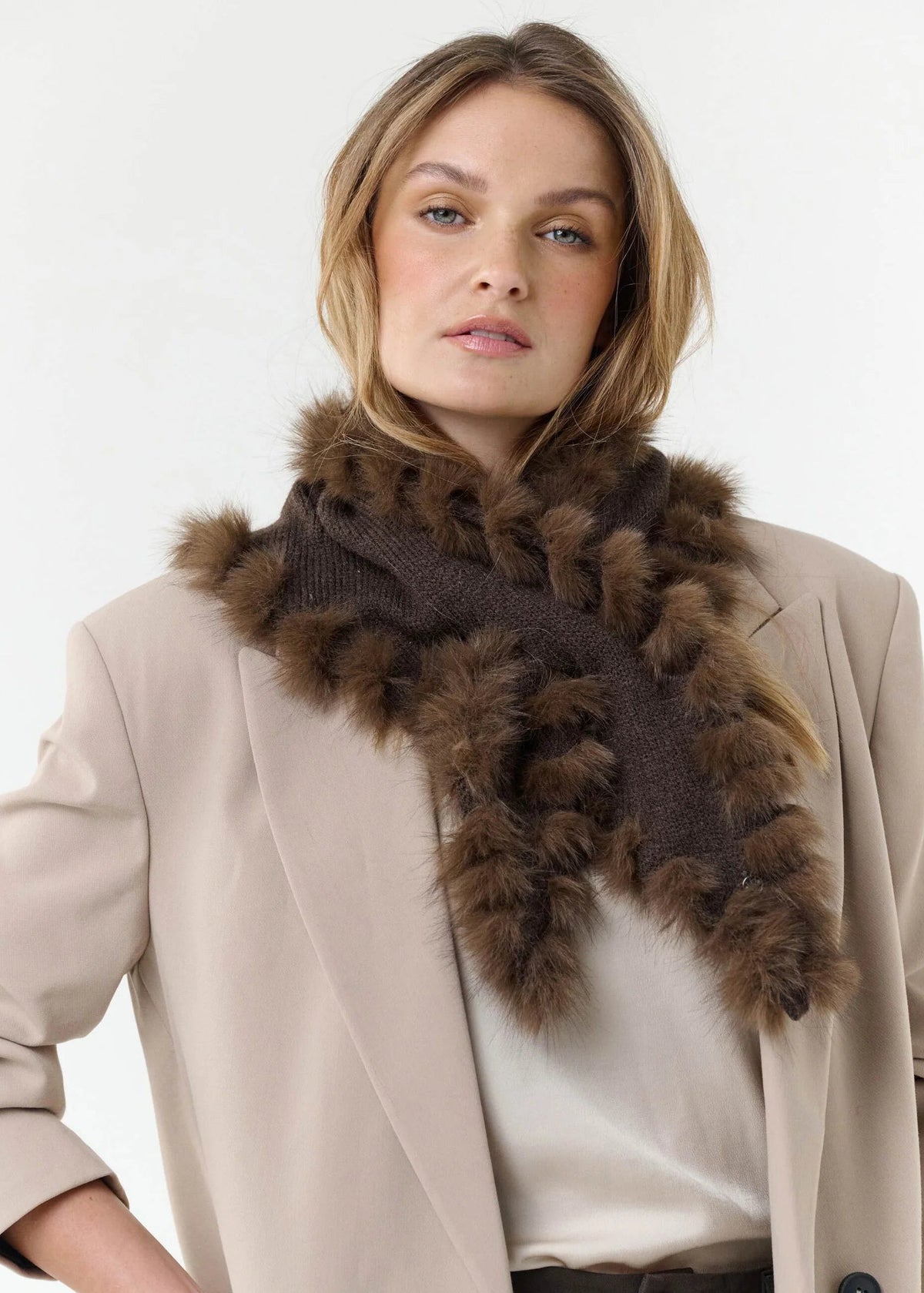 A woman wearing a beige blazer, cream top, and a dark brown knit scarf trimmed with fluffy brown fur, photographed against a plain light background.
