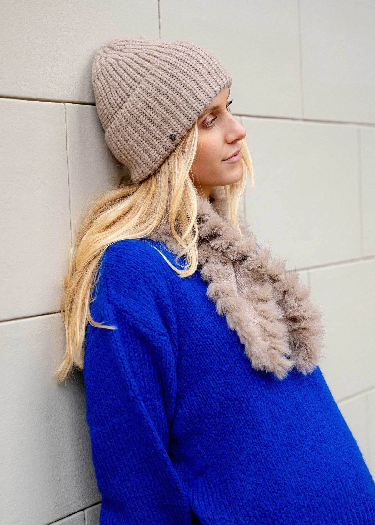 A woman wearing a beige ribbed knit beanie, a fuzzy taupe faux-fur neck warmer, and a bright blue knit sweater leans against a light gray tiled wall.