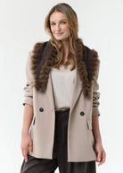 A woman wearing a beige double-breasted coat with a dark brown fur-trimmed collar, styled with a cream top and dark trousers, against a plain white studio background.