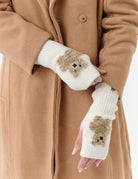 A person wearing a camel-colored coat and white fingerless knit gloves with small brown teddy bear faces on the palms, shown against a plain light background.