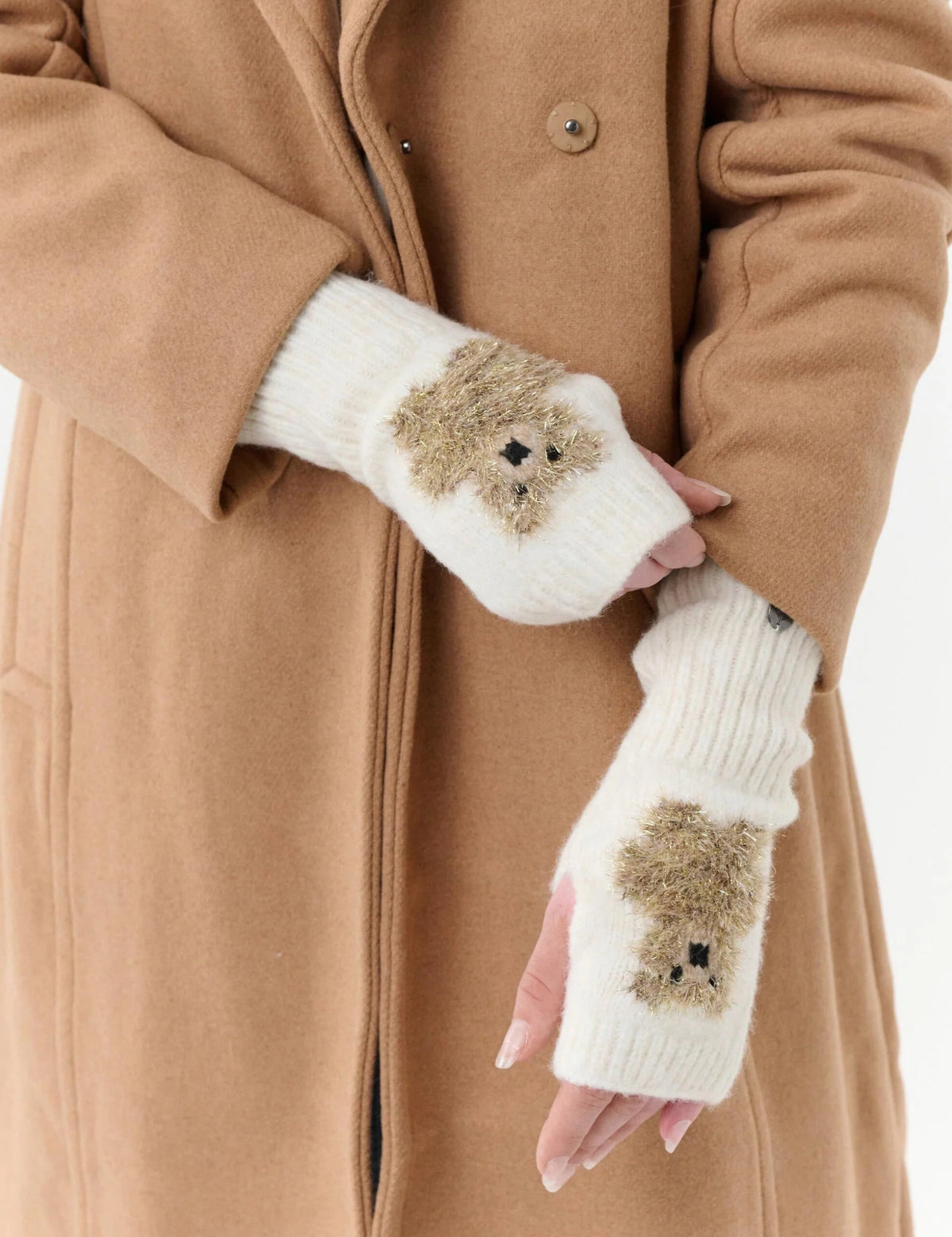 A person wearing a camel-colored coat and white fingerless knit gloves with small brown teddy bear faces on the palms, shown against a plain light background.