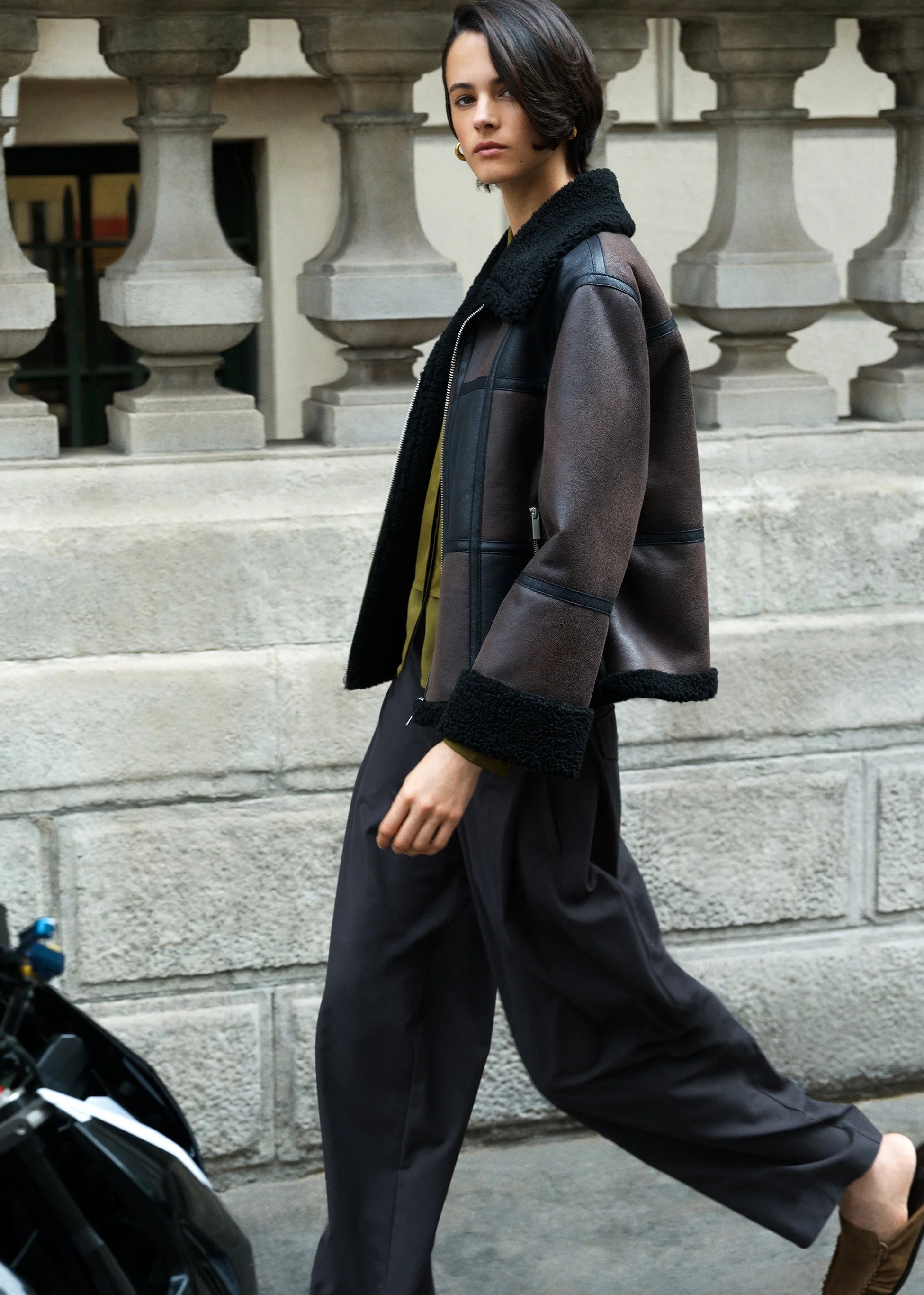 A woman walking outdoors in front of a stone balustrade, wearing a dark brown patchwork shearling jacket with black trim, black wide-leg trousers, and brown loafers.