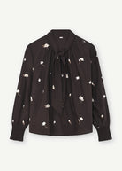 A black long-sleeve blouse with a small white floral print, featuring a tie-neck detail and a ruffled hem, shown against a light off-white background.