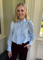 A woman wearing a light blue long-sleeve blouse with ruffled trim down the front and puffed shoulders, paired with dark burgundy velvet trousers, standing indoors near a window.