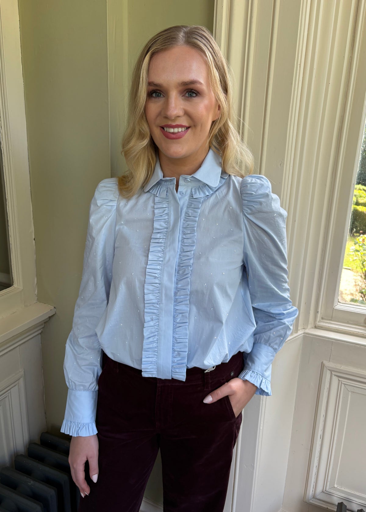 A woman wearing a light blue long-sleeve blouse with ruffled trim down the front and puffed shoulders, paired with dark burgundy velvet trousers, standing indoors near a window.
