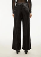 Black wide-leg velvet trousers shown from the back on a model, featuring a textured waistband and back pockets, paired with black heels.