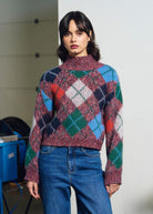 A woman wearing a multicolor argyle knit mock-neck sweater and blue jeans, standing in a minimalist indoor setting with a white wall and some blue equipment in the background.