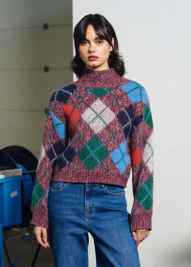 A woman wearing a multicolor argyle knit mock-neck sweater and blue jeans, standing in a minimalist indoor setting with a white wall and some blue equipment in the background.