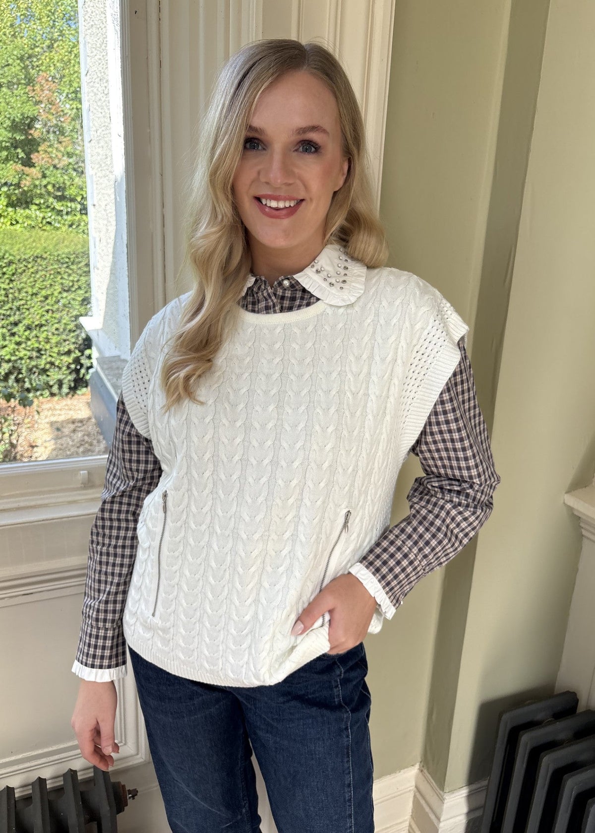 A woman wearing a cream cable-knit sleeveless vest over a black and white gingham shirt with blue jeans, standing in a room with a window and light green walls.